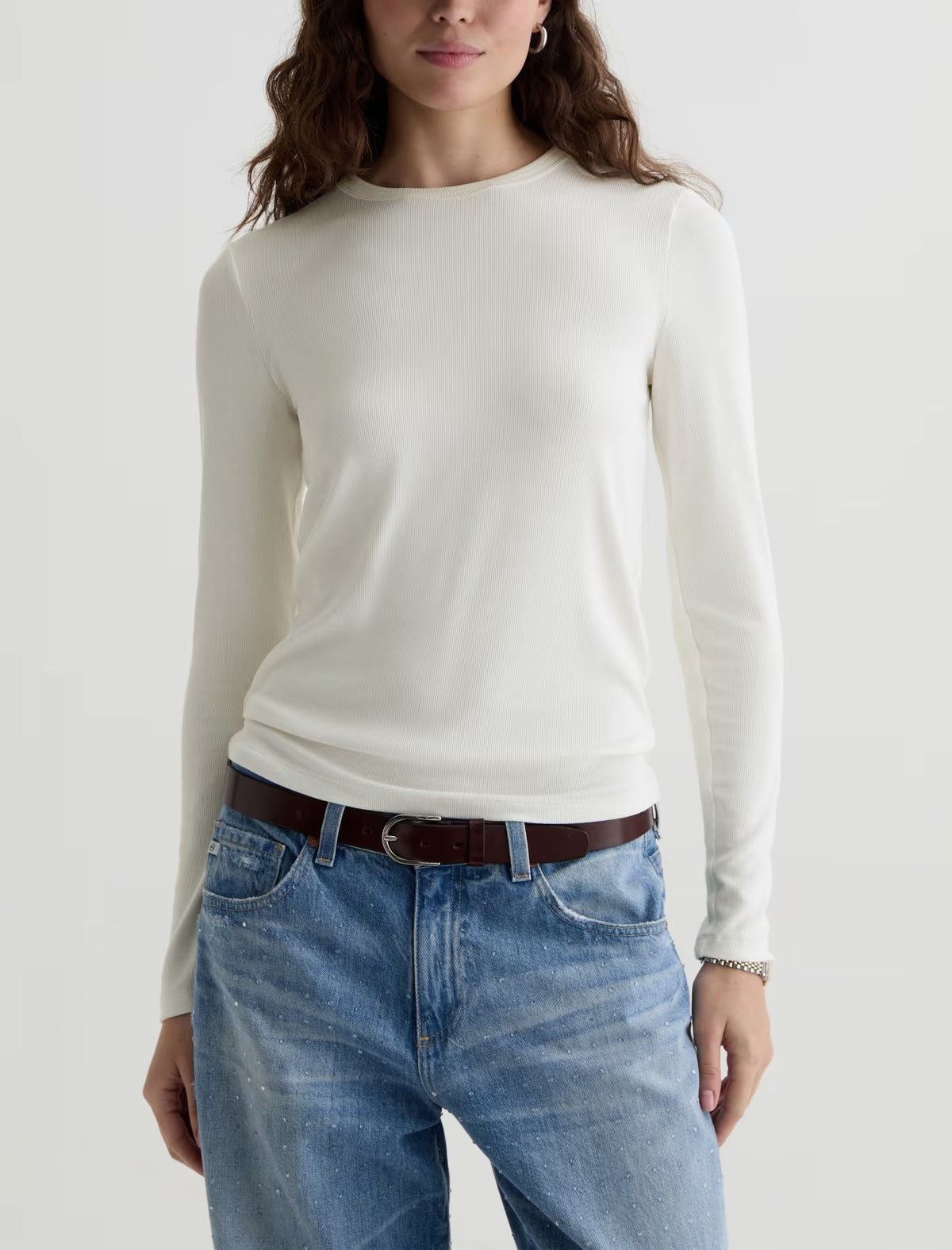 A woman wearing a fitted off-white long-sleeve knit top tucked into light-wash blue jeans with a brown belt, photographed against a plain light gray studio background.