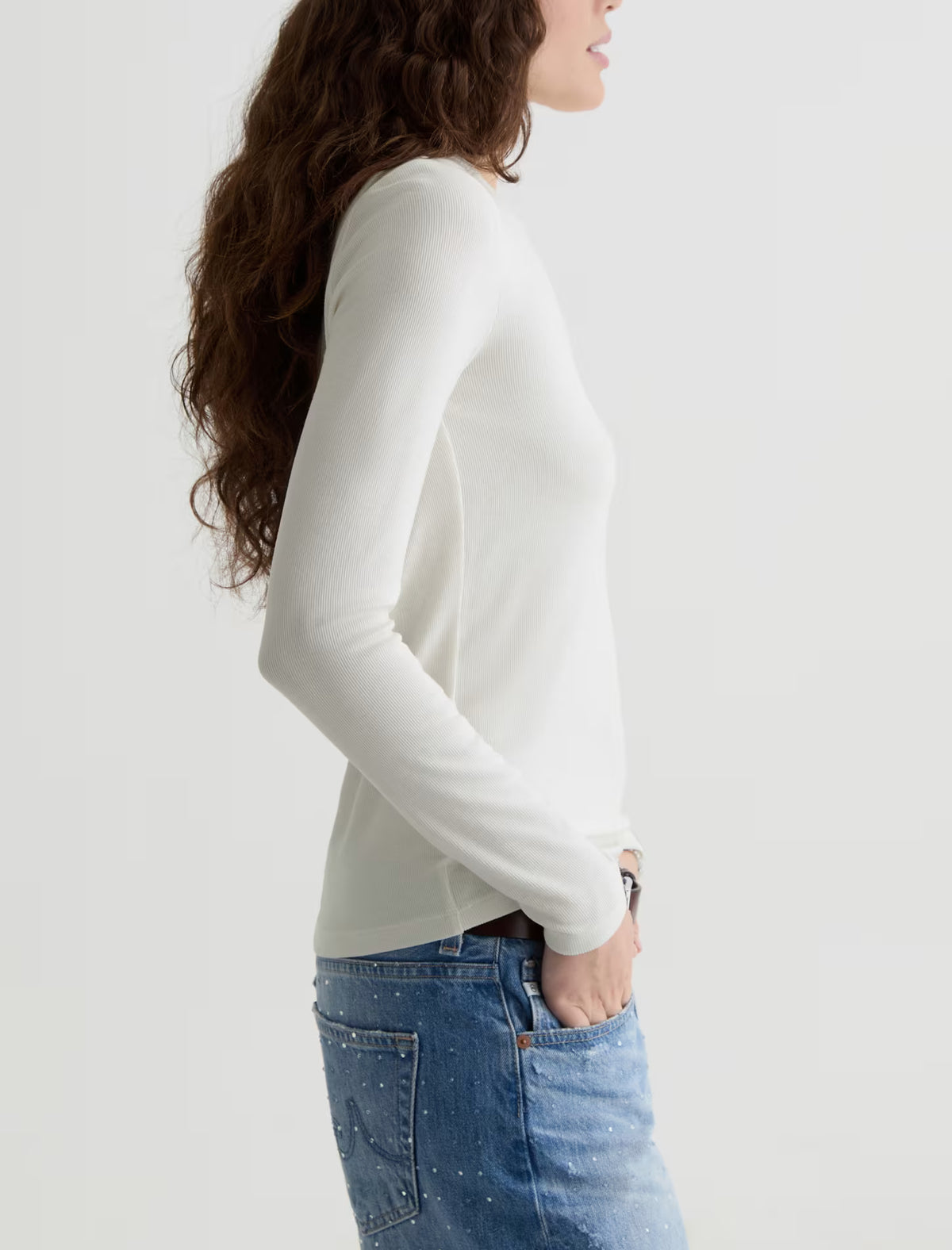 A woman shown in side profile wearing a fitted long-sleeve white top and blue jeans against a plain light background.