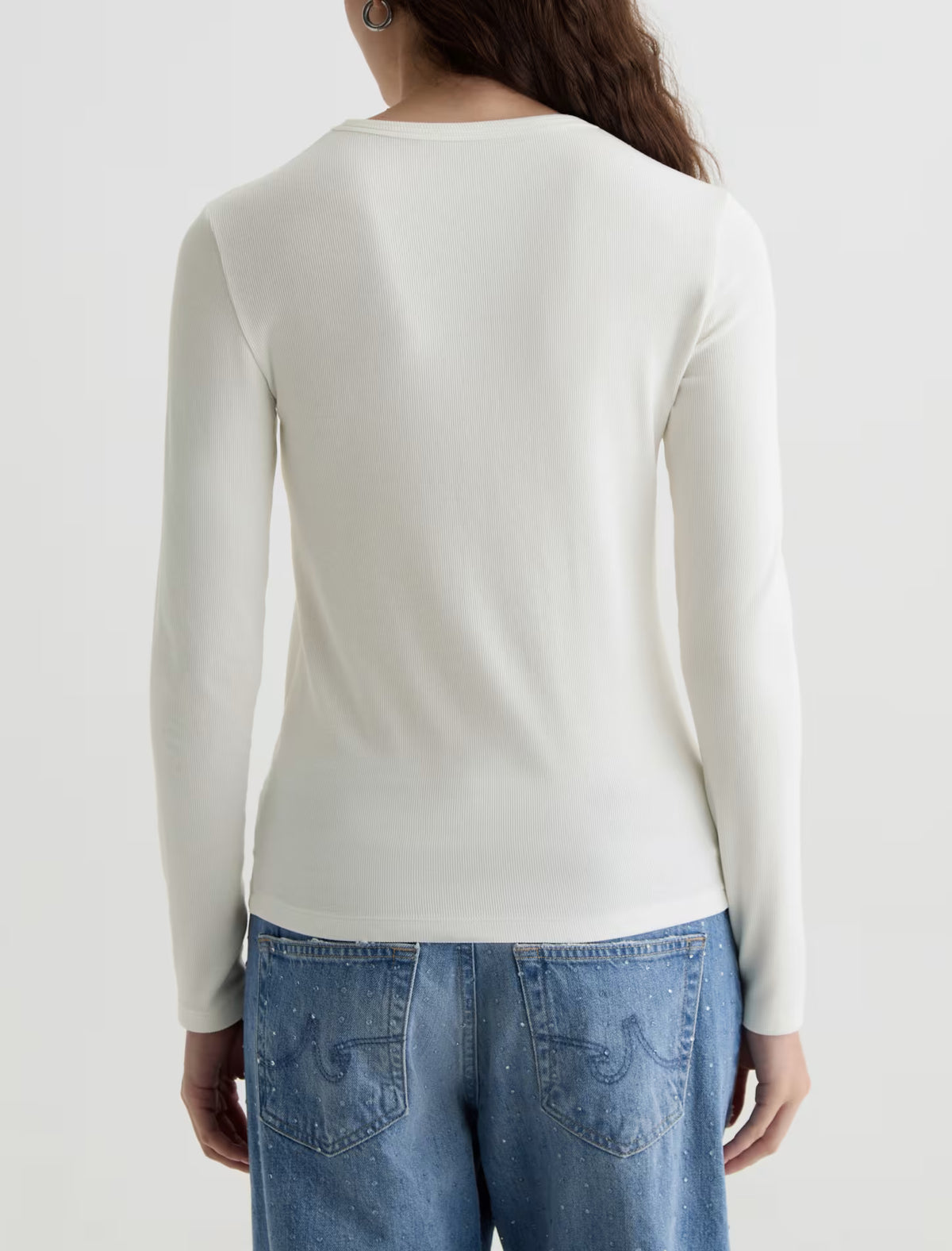 A woman viewed from the back wearing a plain off-white long-sleeve knit top and blue jeans against a clean light gray studio background.