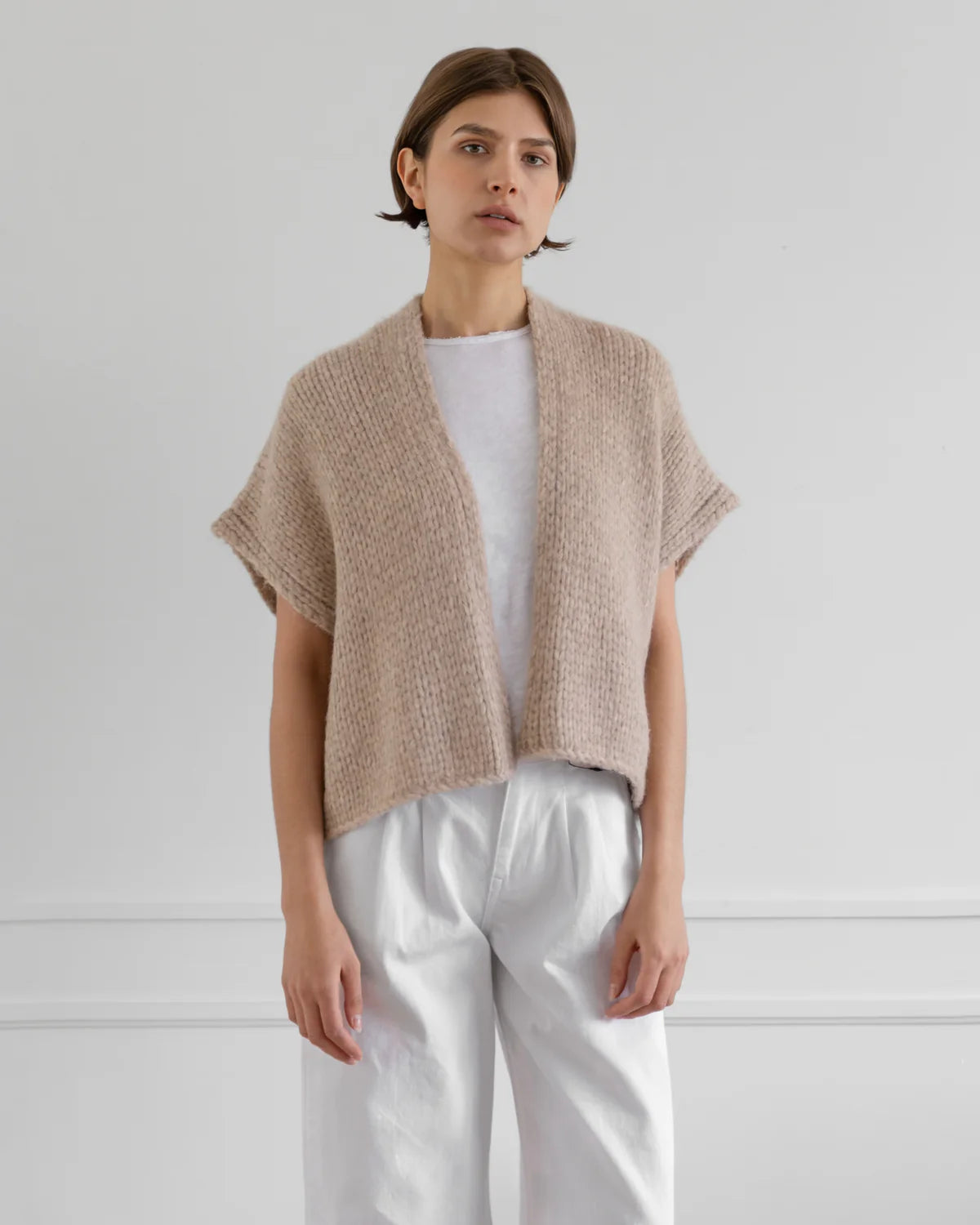 Woman wearing a beige knitted cardigan over a white shirt and pants on a light gray background