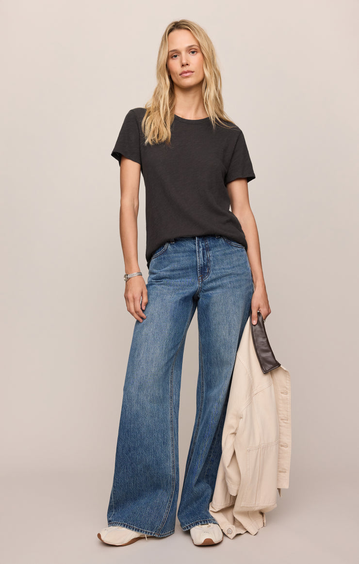 Woman wearing a black t-shirt and blue jeans holding a beige jacket against a neutral background