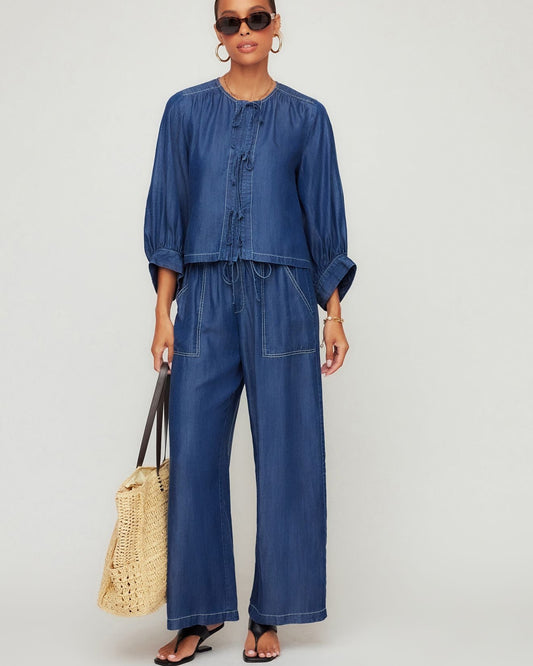 Woman wearing a blue denim outfit with sunglasses and a straw bag on a plain background