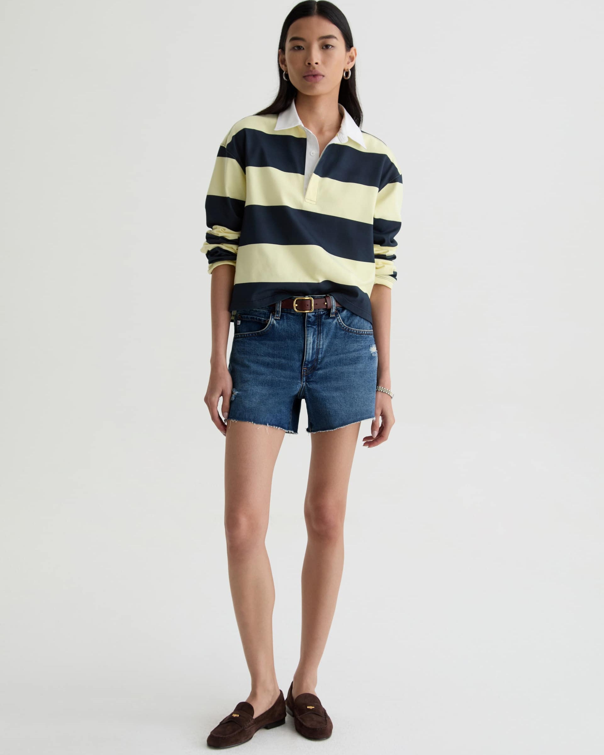 Woman wearing a striped rugby shirt, denim shorts, and brown loafers on a white background
