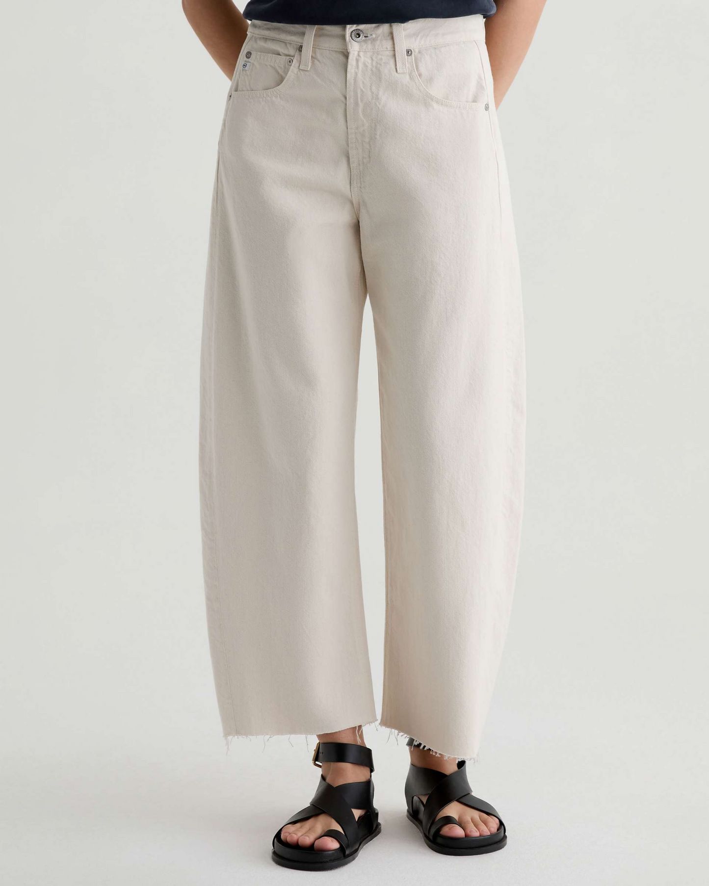 A person wearing off-white wide-leg cropped jeans with a raw hem and black slide sandals, shown from the waist down against a plain light gray studio background.