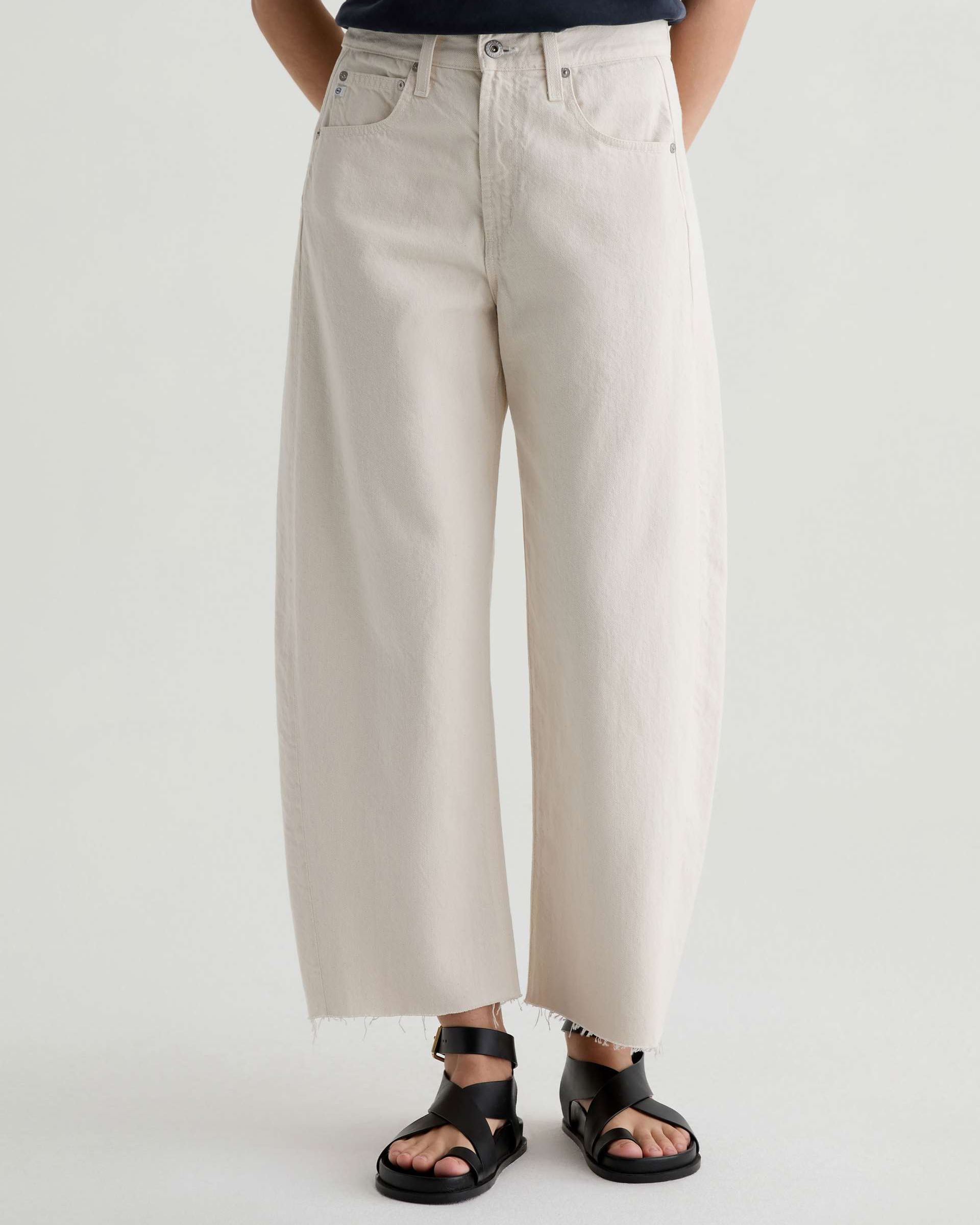 A person wearing off-white wide-leg cropped jeans with a raw hem and black slide sandals, shown from the waist down against a plain light gray studio background.