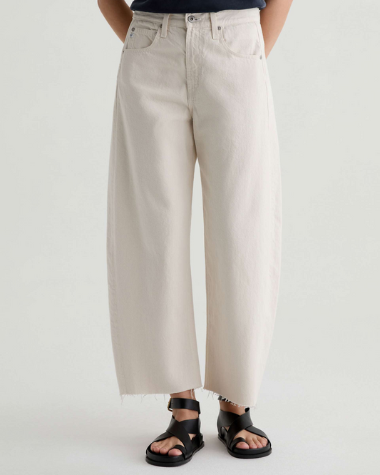 A person wearing off-white wide-leg cropped jeans with a raw hem and black slide sandals, shown from the waist down against a plain light gray studio background.