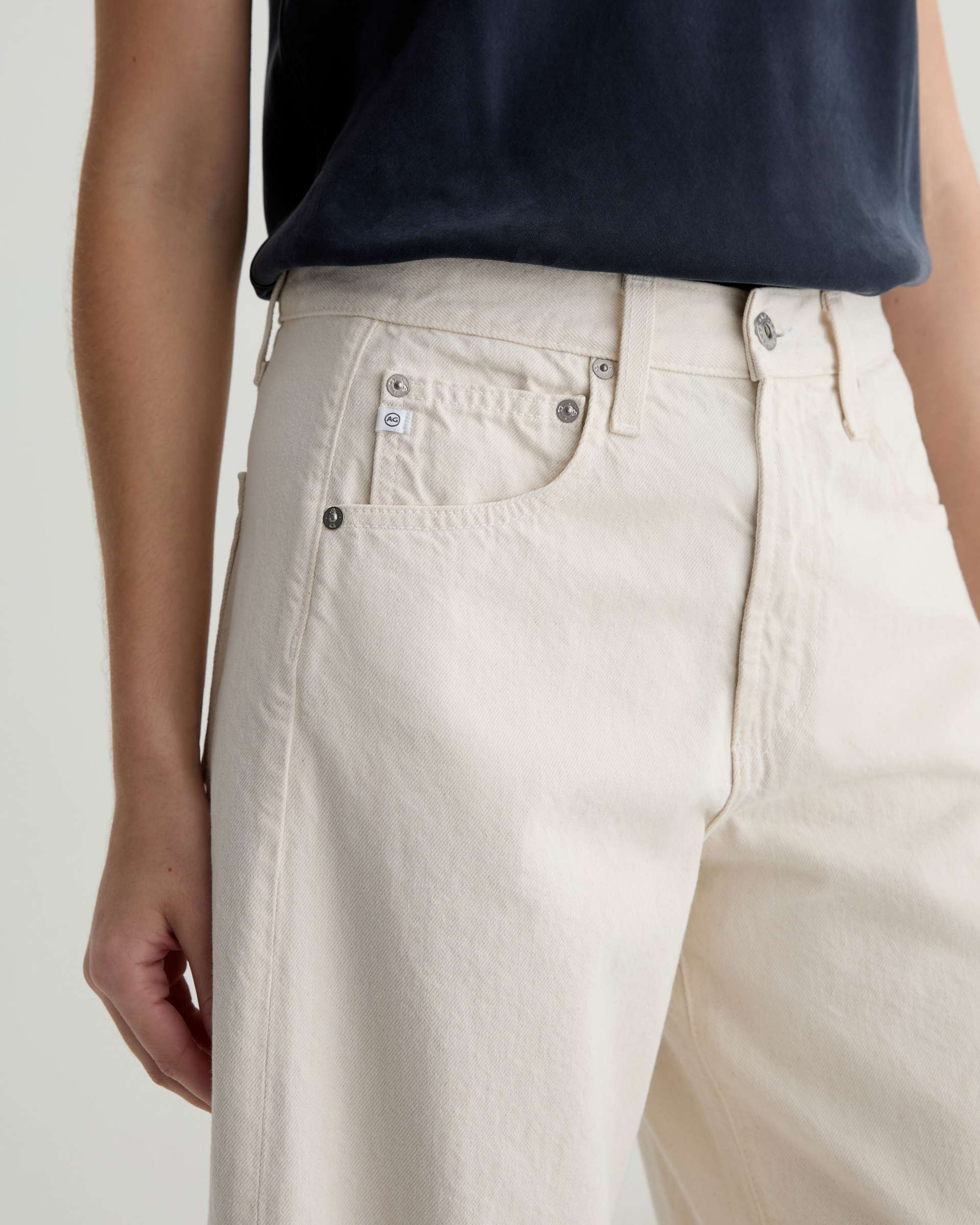 Close-up of a person wearing light beige wide-leg denim jeans with a navy sleeveless top, showing the front pocket and waistband details.