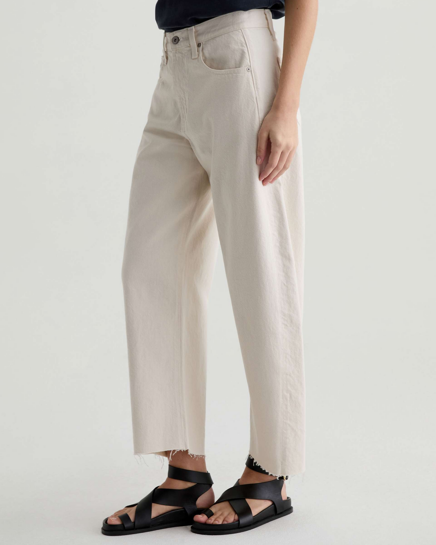 A person wearing high-waisted, wide-leg cropped pants in a light beige or cream tone, paired with black strappy sandals against a plain light gray studio background.