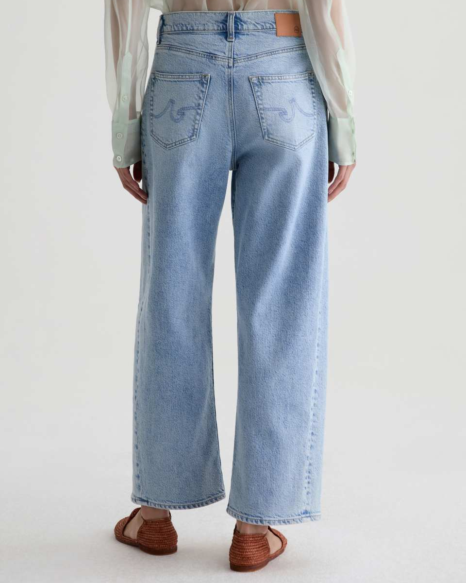 A person shown from the back wearing light-wash wide-leg jeans with embroidered details on the back pockets, a sheer pale green blouse, and brown wedge sandals against a plain light studio background.
