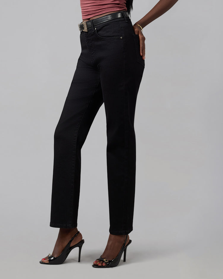 A model wearing black straight-leg jeans, styled with a black leather belt, a pink top, and black high-heeled sandals, photographed against a neutral light gray studio background.