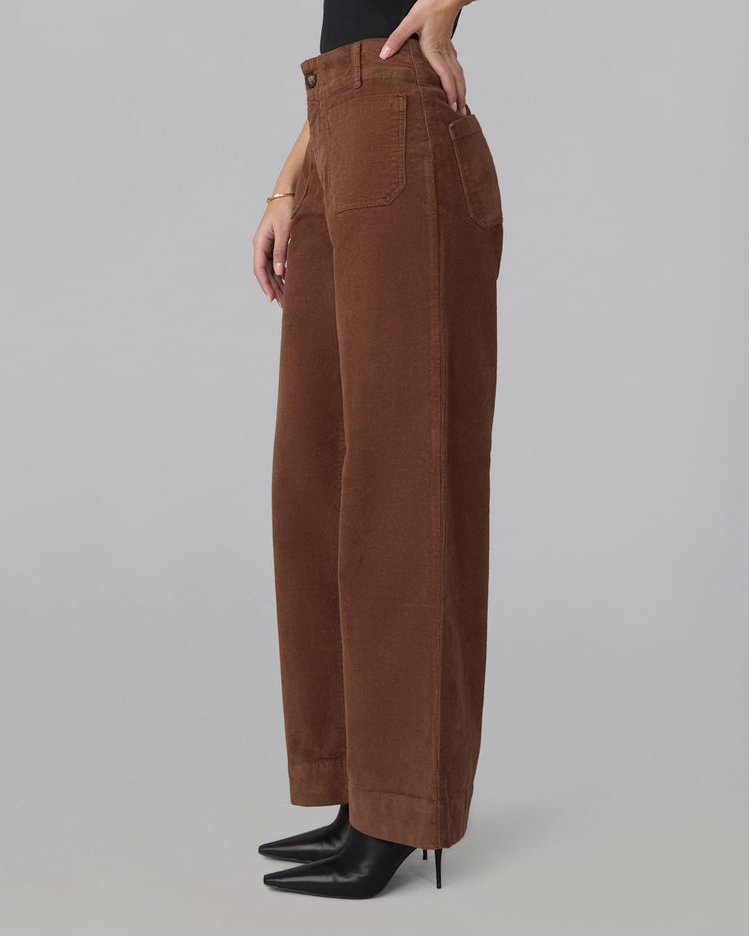 A person wearing high-waisted, wide-leg brown corduroy pants with black pointed-toe ankle boots, shown from the waist down against a plain light gray studio background.