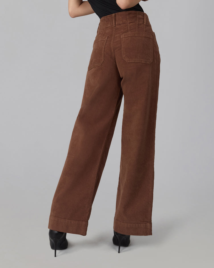 A person shown from the back wearing high-waisted, wide-leg brown corduroy pants with black high heels against a plain light gray studio background.