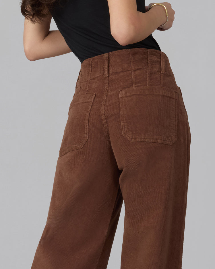 A close-up back view of a person wearing high-waisted brown corduroy pants with a black top, set against a solid gray studio background.