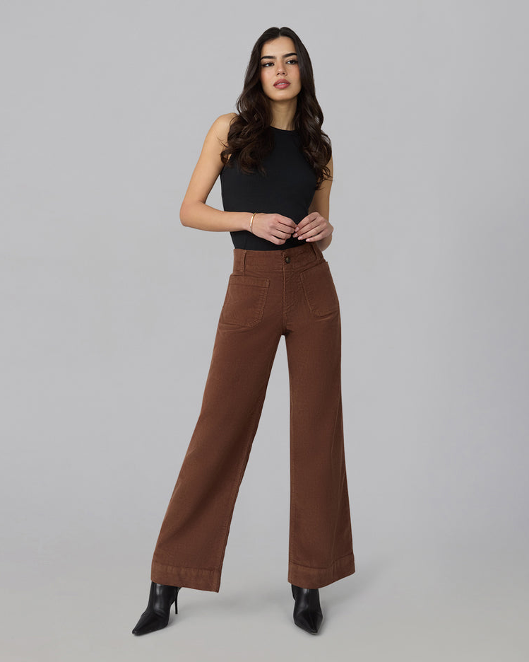 A woman modeling a pair of high-waisted, wide-leg brown pants with a black sleeveless top and black pointed-toe heels, standing against a solid light gray studio background.