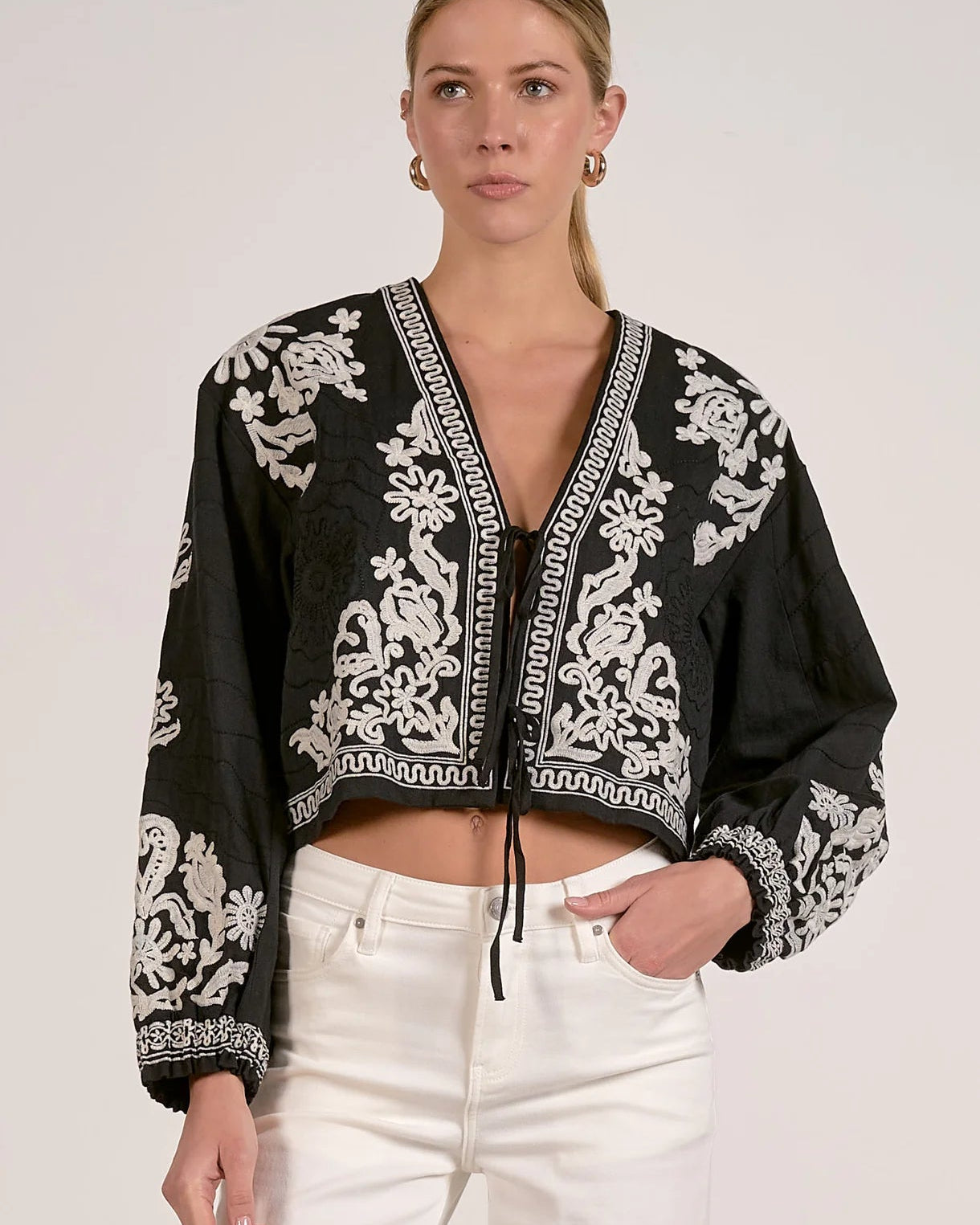 Woman wearing a black and white embroidered top with white pants on a plain background