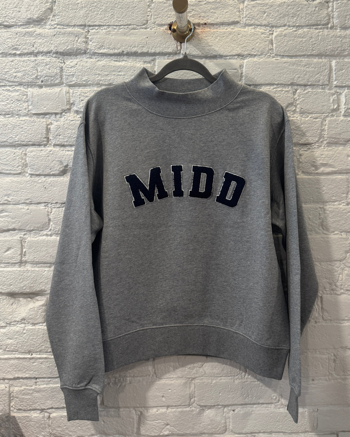 A heather gray crewneck sweatshirt with the word 'MIDD' in dark navy blue varsity-style lettering across the chest, hanging on a black hanger against a white painted brick wall.