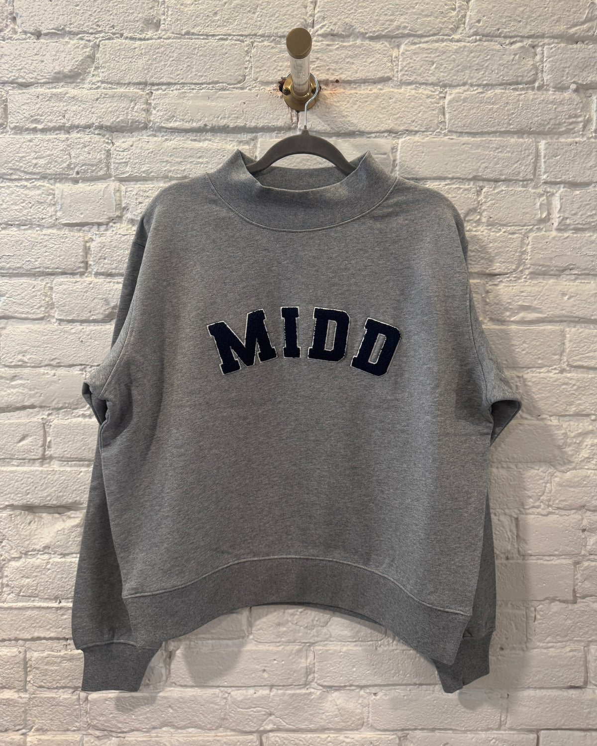A heather gray cropped sweatshirt with 'MIDD' embroidered in navy blue varsity-style letters across the chest, hanging on a black hanger against a white painted brick wall.