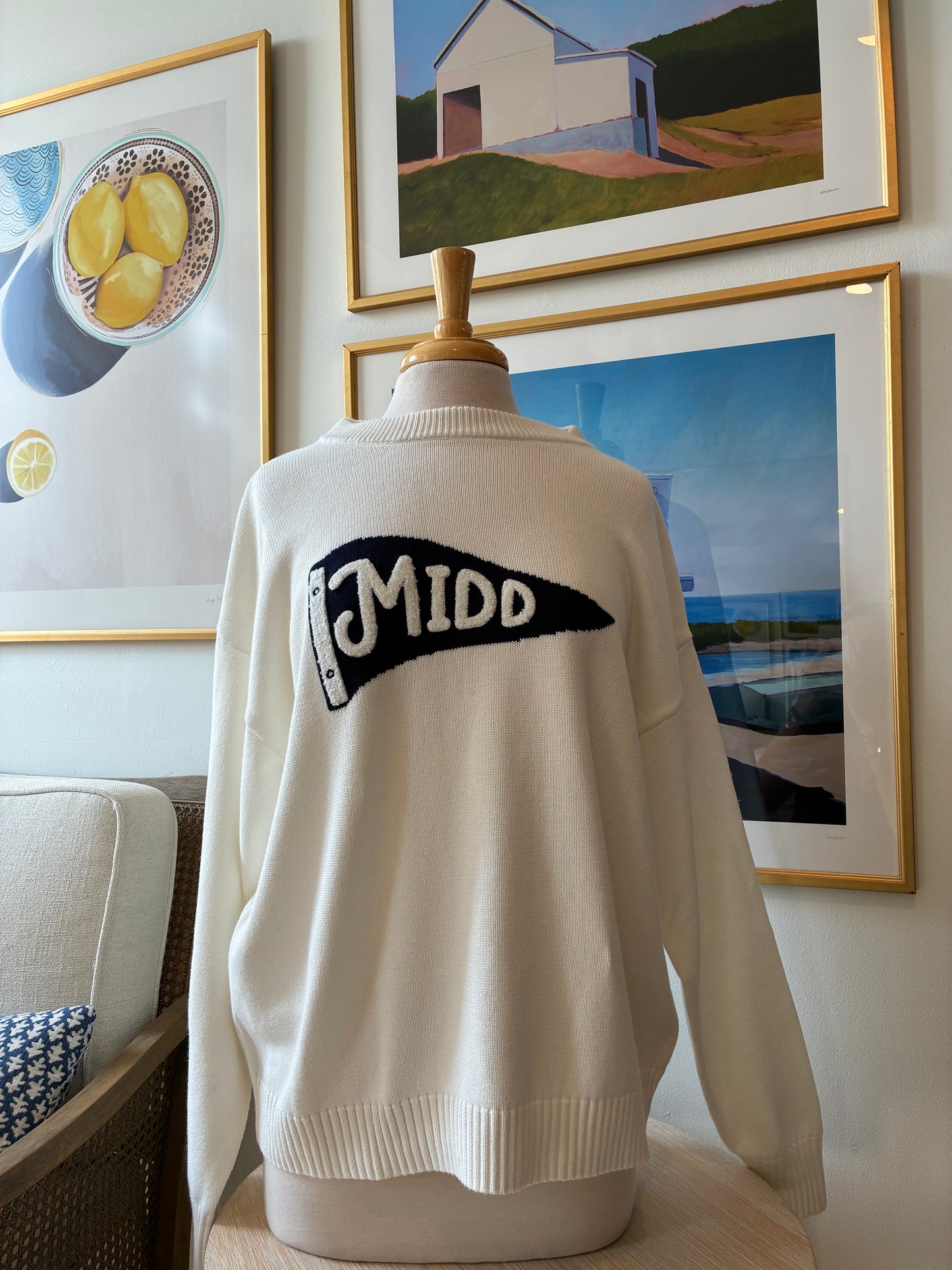 A cream-colored knit sweater displayed on a mannequin torso in an indoor room setting, featuring a black and white 'MIDD' pennant graphic on the back.