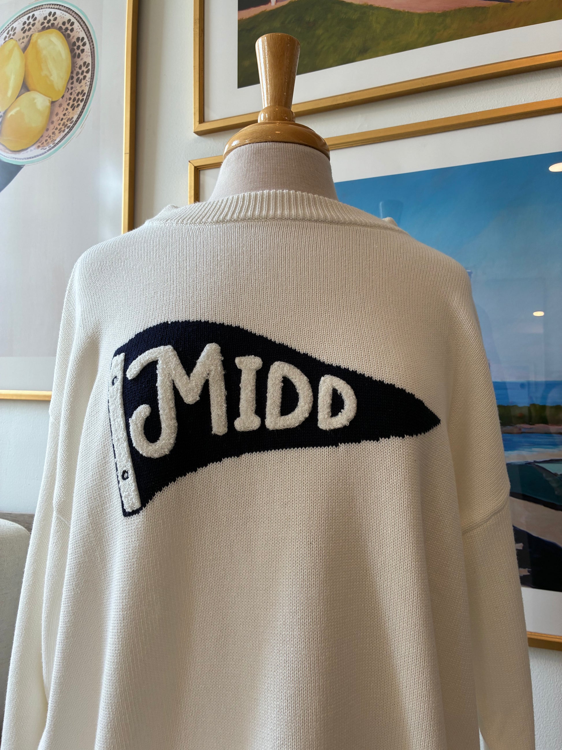 An off-white knit sweater displayed on a mannequin torso, featuring a large black pennant-shaped graphic with the word 'MIDD' in white lettering across the chest.