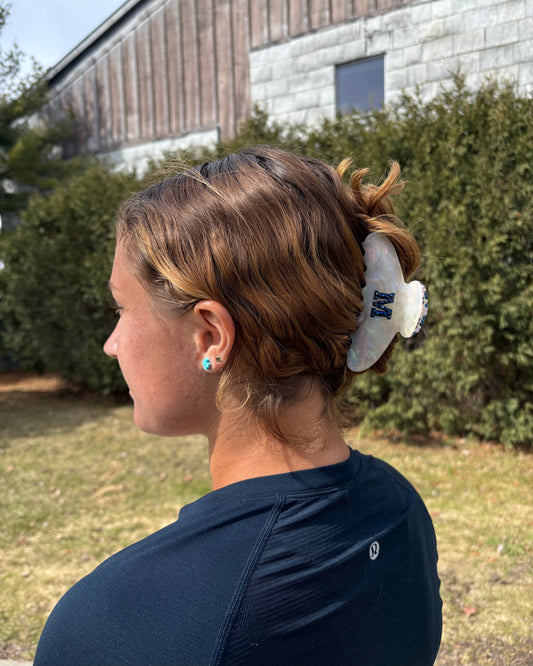 Person with brown hairwearing a M hairclip outside with grass and trees.