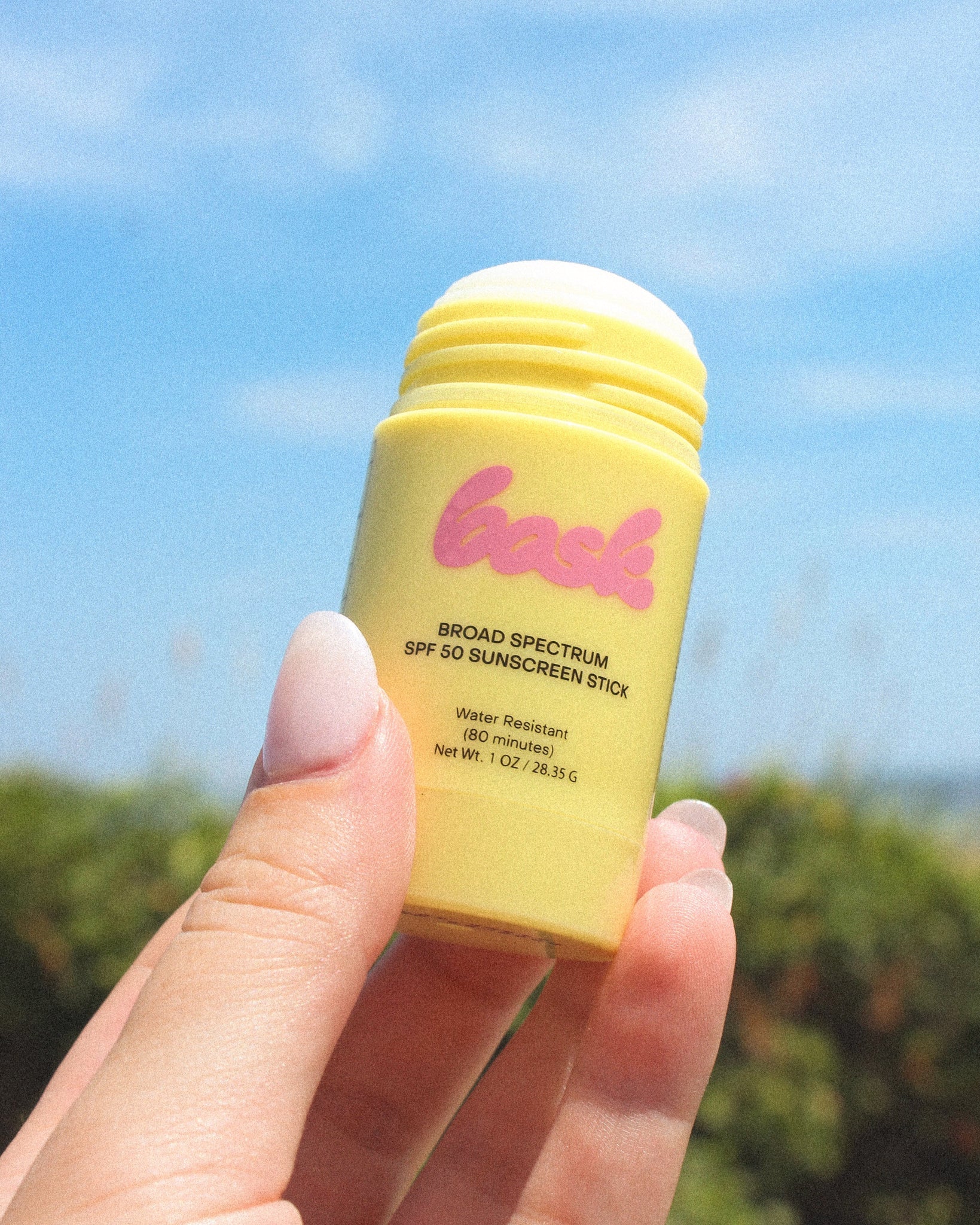 Yellow sunscreen stick with pink logo held in a hand against a blue sky.