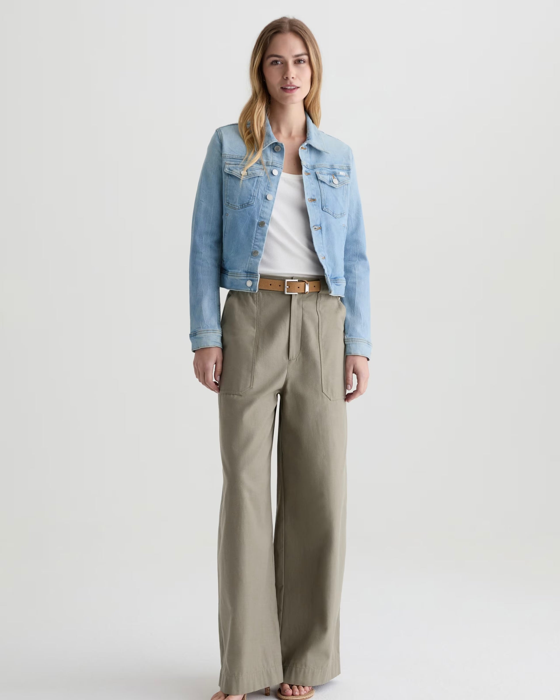 Woman wearing a denim jacket, white shirt, and beige pants on a light gray background