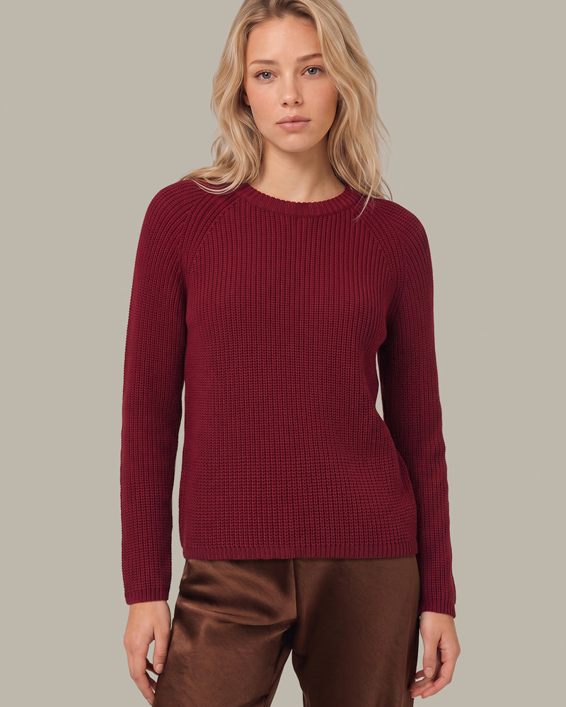 A woman modeling a long-sleeve ribbed knit sweater in deep burgundy, paired with brown satin pants, against a solid light gray background.