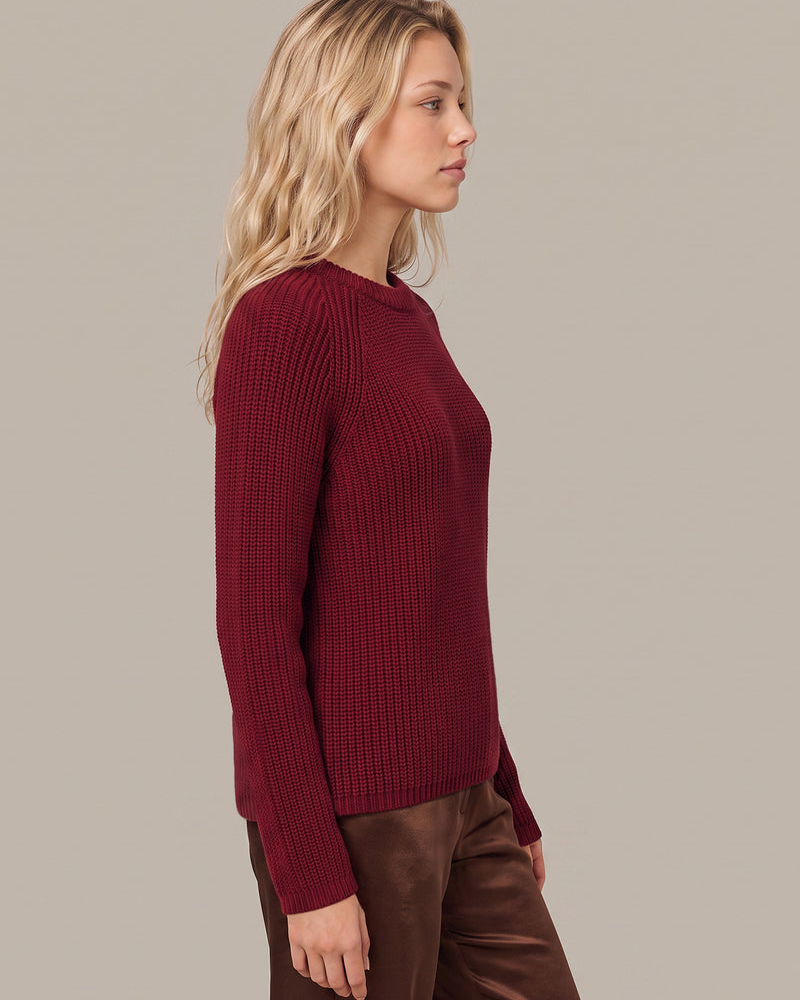 A woman in profile wearing a ribbed long-sleeve crewneck sweater in deep burgundy, paired with brown pants, against a solid light gray studio background.