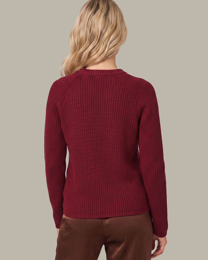 A woman viewed from the back wearing a ribbed knit long-sleeve sweater in deep burgundy and brown pants, set against a solid light gray background.