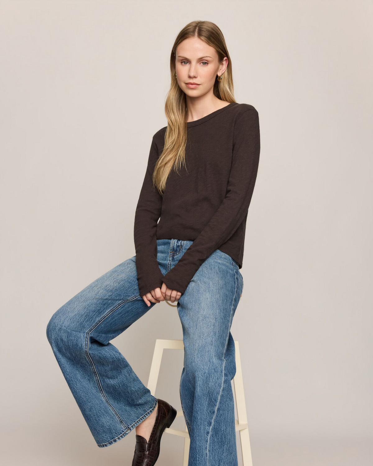 A woman sits on a white stool wearing a dark brown long-sleeve knit top, blue wide-leg jeans, and dark brown leather loafers against a light gray studio background.