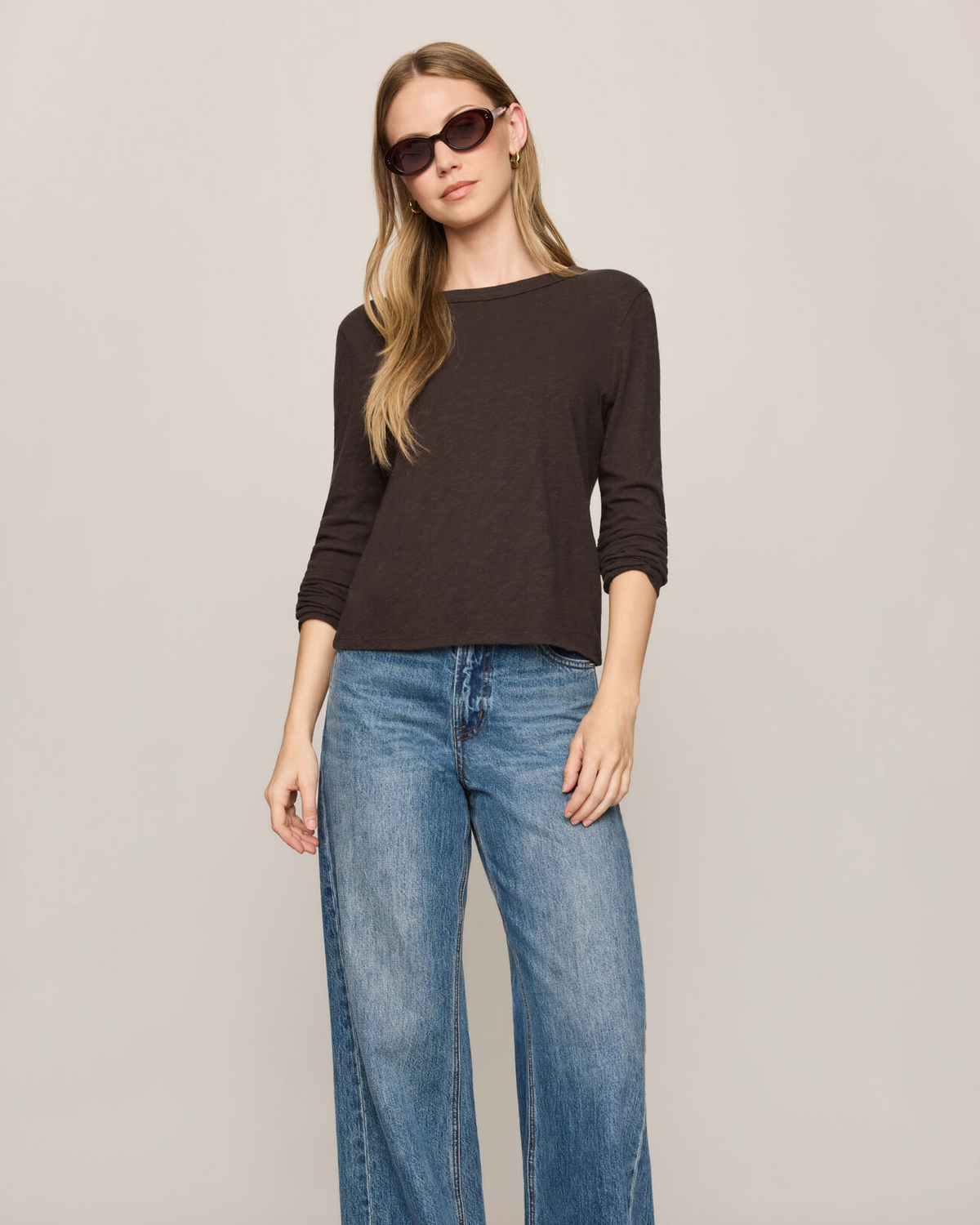 A woman modeling a dark brown long-sleeve knit top and wide-leg blue jeans against a plain light gray studio background.