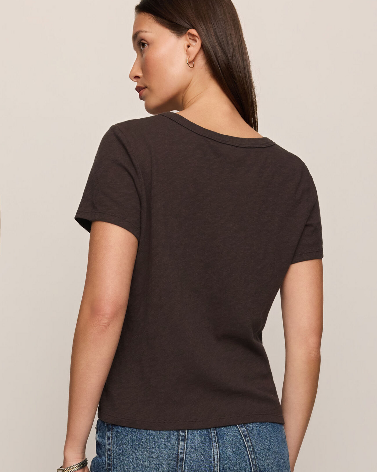 A woman shown from the back wearing a dark brown short-sleeve crewneck t-shirt and blue jeans, photographed against a solid light beige background.