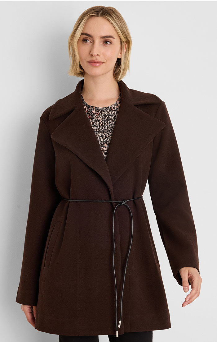A woman wearing a dark brown wrap-style coat with a thin black tie belt, layered over a patterned top, photographed against a light gray studio background.
