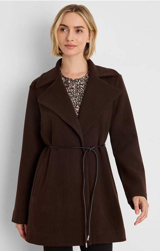 A woman wearing a dark brown wrap-style coat with a thin black tie belt, layered over a patterned top, photographed against a light gray studio background.