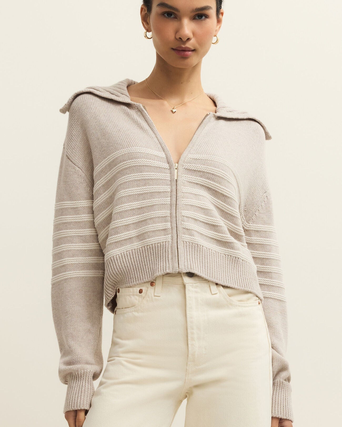 Woman wearing a light gray zip-up sweater with white pants on a beige background