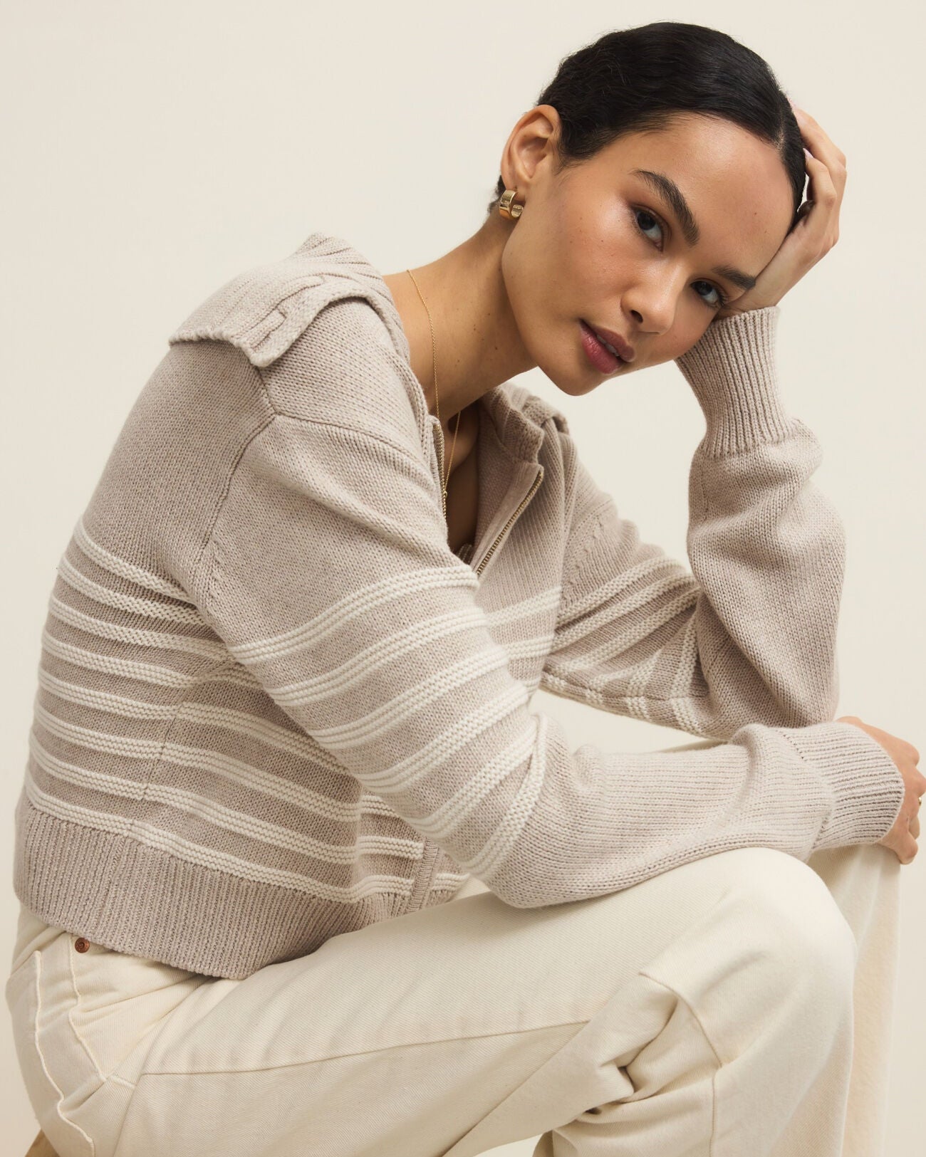 Woman wearing a beige striped sweater and white pants sitting on a wooden stool.
