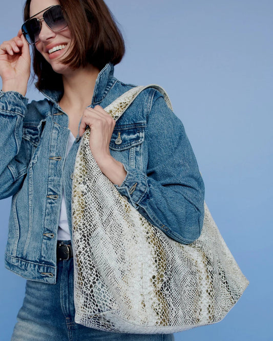Woman wearing a denim jacket and sunglasses, holding a patterned bag against a blue background
