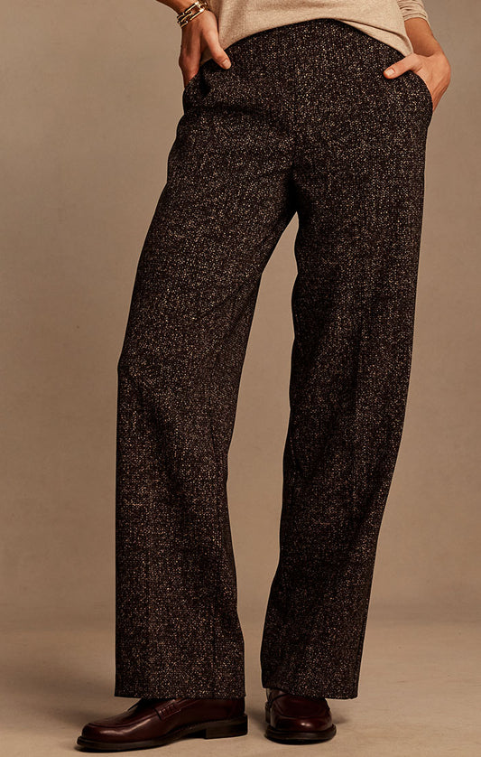 A person wearing dark brown and black textured tweed trousers with a beige top and dark brown leather dress shoes, standing against a solid beige studio background.