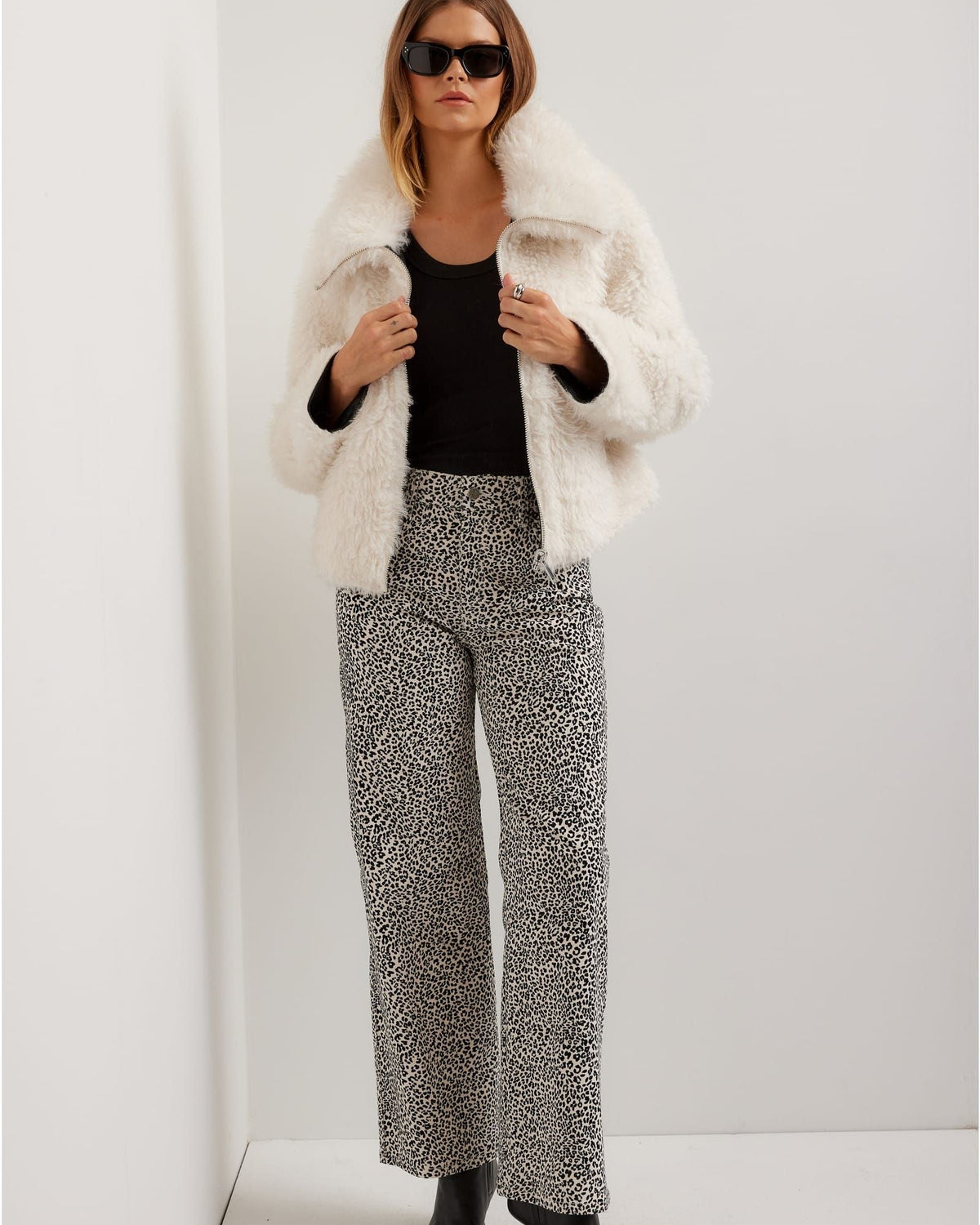 Woman wearing a white fur coat, black top, and patterned pants against a white background