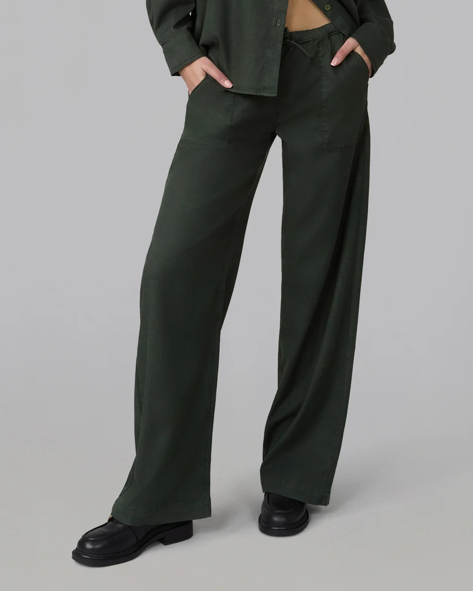 A model wearing dark olive green wide-leg trousers with a matching dark green button-up shirt and black leather shoes, standing against a solid gray studio background.