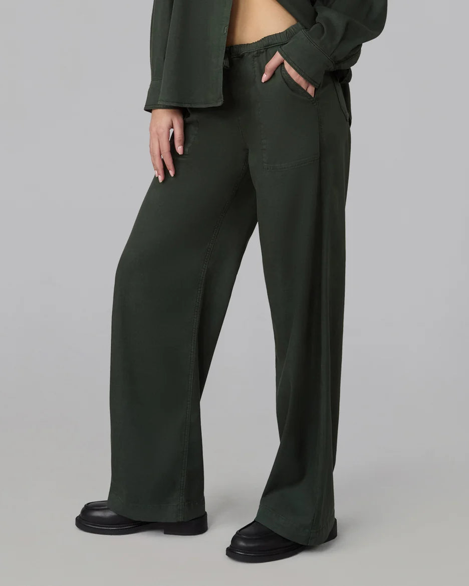 A model wearing dark olive green wide-leg trousers with a drawstring waist and side pockets, styled with a matching dark green cropped top and black leather shoes against a neutral gray studio background.