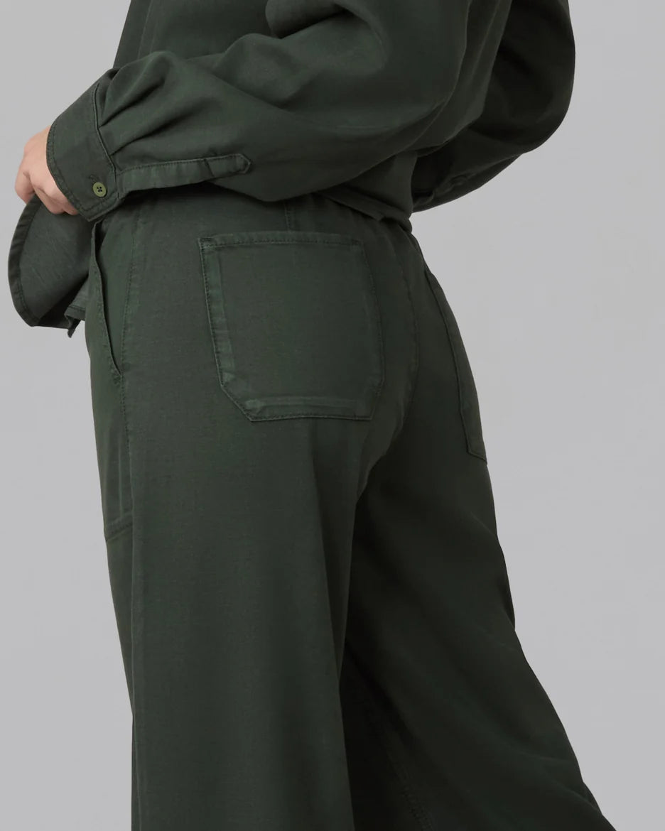 Close-up side view of a person wearing dark olive green wide-leg trousers with a back pocket, paired with a matching long-sleeve top, against a plain light gray studio background.