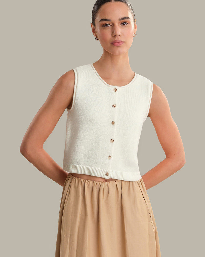 Woman wearing a white sleeveless knit top with buttons and a beige skirt on a neutral background