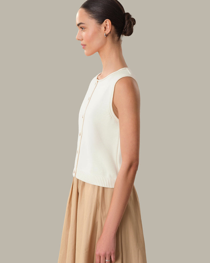 Woman wearing a white sleeveless top and beige skirt against a neutral background