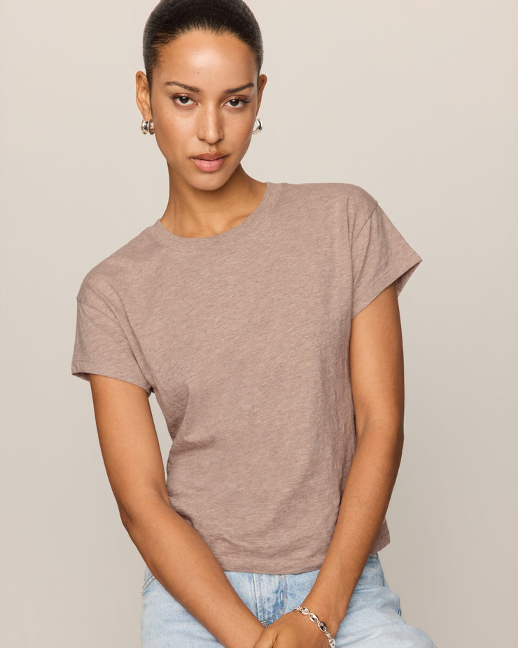 A woman wearing a short-sleeve crewneck t-shirt in a heathered taupe-brown color, paired with light-wash blue jeans, posed against a solid light beige background.