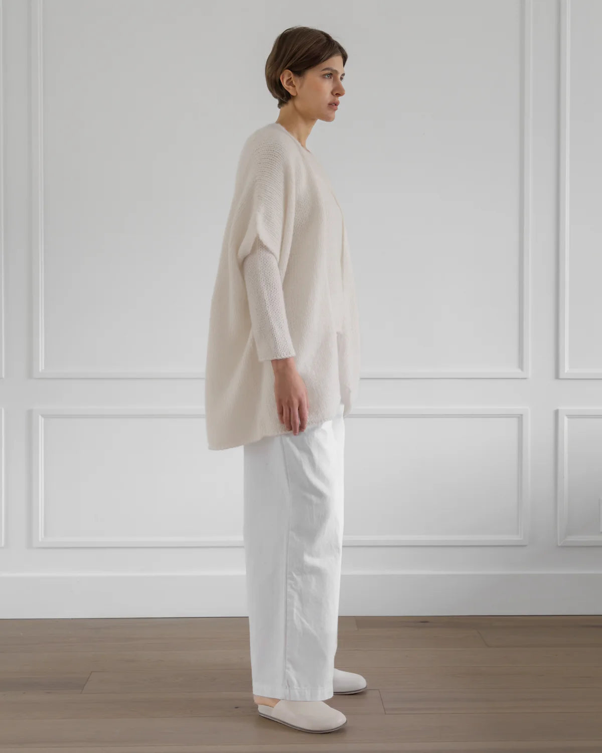 Woman wearing a beige cardigan and white pants standing against a white paneled wall.