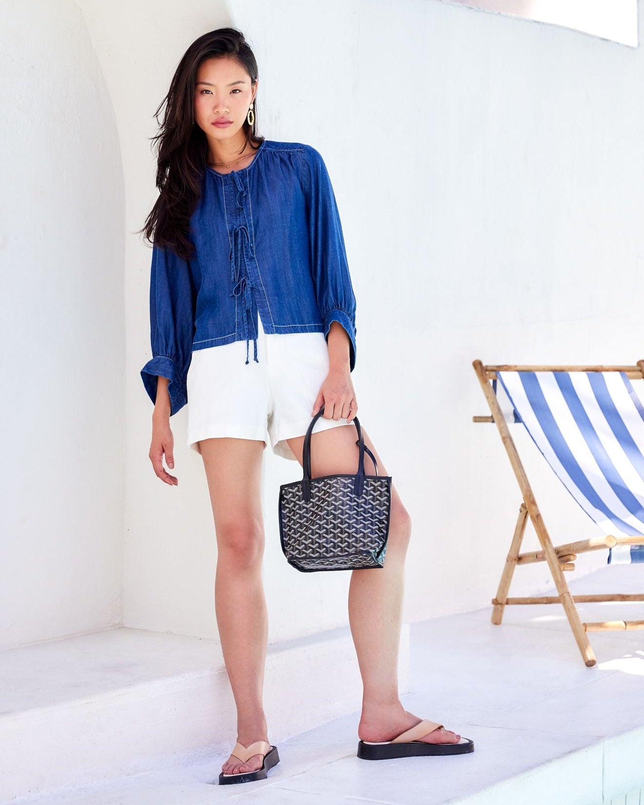 Woman in blue shirt and white shorts holding a black handbag in a bright room with a striped chair.