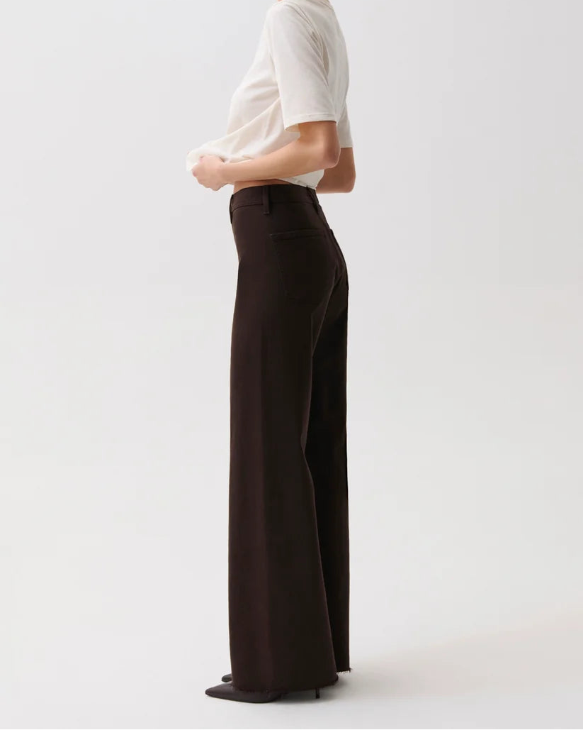 A woman stands in profile wearing a fitted white short-sleeve top and dark brown wide-leg trousers against a clean light gray studio background.