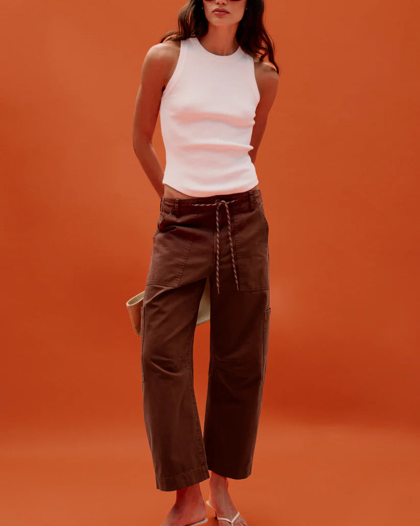 A model stands against a solid orange studio background wearing a white ribbed sleeveless top, dark brown wide-leg cropped pants with a drawstring waist, and clear thong sandals.
