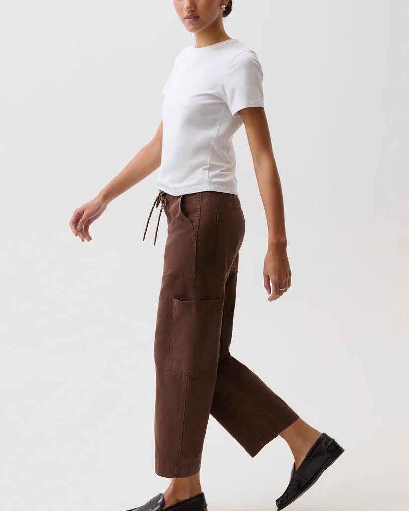 A woman modeling a pair of dark brown cropped trousers with a drawstring waist, styled with a white t-shirt and black loafers against a plain light gray studio background.